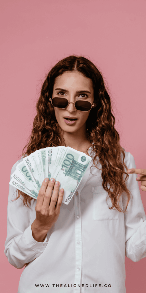woman holding a handful of cash in front of a pink wall | Signs That Money Is Coming Your Way | The Aligned Life Manifestation Blog